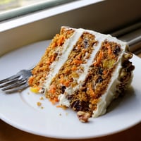 A slice of spiced Carrot Cake with cream cheese frosting and a toasted walnut garnish on a white plate.