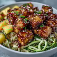 Golden-brown baked tofu cubes and spiralized zucchini and carrots sit atop fluffy quinoa in a white bowl.